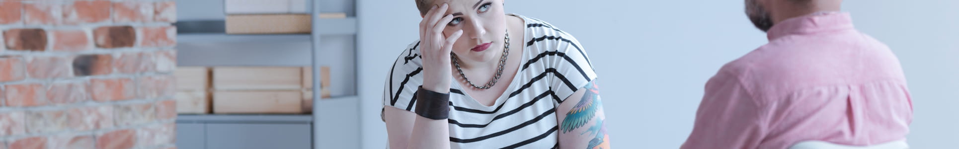 Young rebellious woman sadly propping head up on her arm rest on her knee while speaking with a therapist